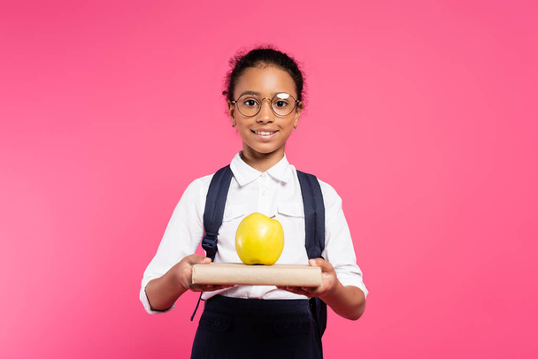 smiling african american schoolgirl in glasses with book and apple isolated on pink - Фото, изображение