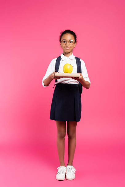 smiling african american schoolgirl in glasses with book and apple on pink - Foto, imagen
