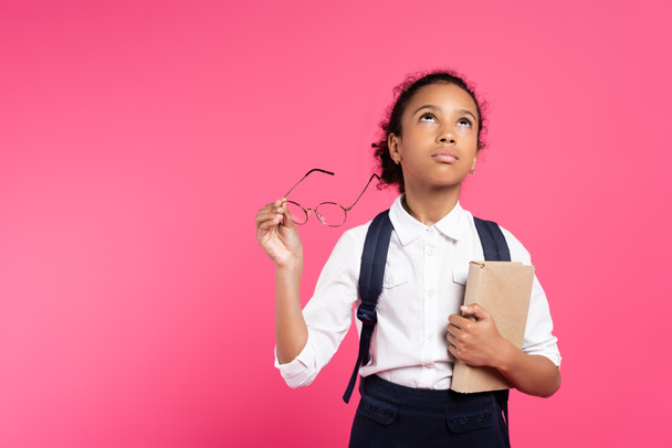 duur Afrikaans amerikaans schoolmeisje met boek en bril op zoek omhoog geïsoleerd op roze - Foto, afbeelding