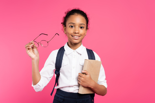 mosolygós afro-amerikai iskolás lány könyv és szemüveg elszigetelt rózsaszín - Fotó, kép