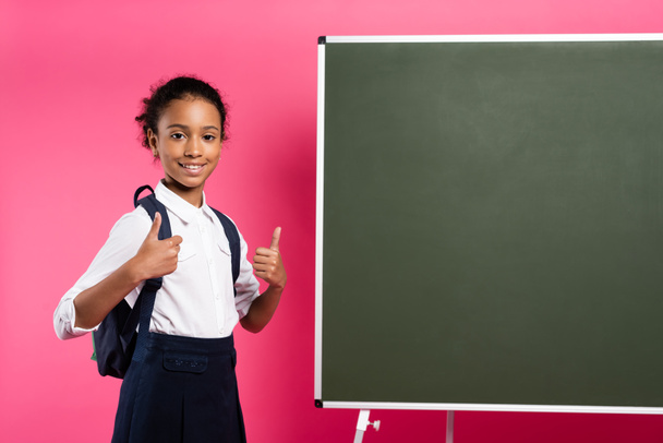 улыбающаяся африканская школьница с рюкзаком рядом с пустой доской, показывающая большие пальцы на розовом фоне
 - Фото, изображение