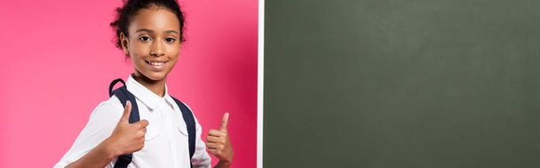 smiling african american schoolgirl with backpack near empty chalkboard showing thumbs up on pink background, panoramic shot - Photo, Image