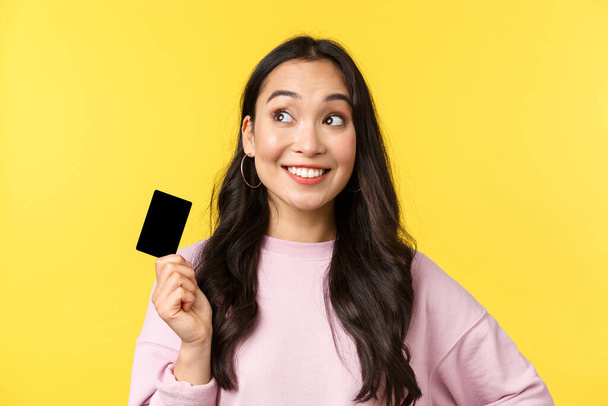 People emotions, lifestyle leisure and beauty concept. Stylish cute asian girl dreaming of new clothes for summer trip, holding credit card and looking upper left corner thoughtful, smiling - Foto, afbeelding