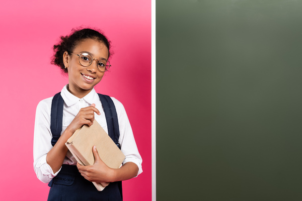 smiling african american schoolgirl with book near empty chalkboard on pink background - Photo, Image