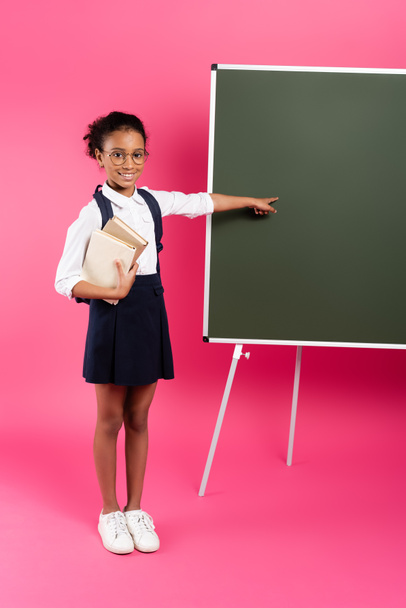 smiling african american schoolgirl with books pointing at empty chalkboard on pink background - Fotó, kép
