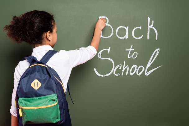 vue arrière de l'écolière afro-américaine avec sac à dos écrivant inscription retour à l'école sur tableau vert - Photo, image