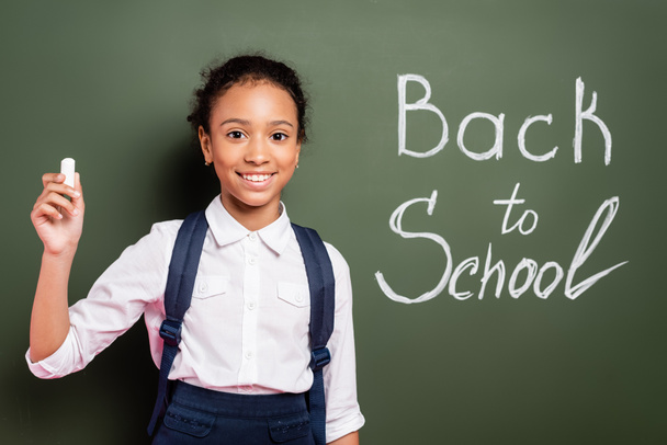 smiling african american schoolgirl with chalk near back to school inscription on green chalkboard - Foto, Bild