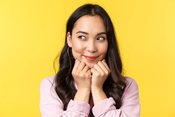 People emotions, lifestyle leisure and beauty concept. Silly dreamy asian girl have interesting idea in mind, imaging something as peeking left and smiling, scheming plan, yellow background - Photo, image
