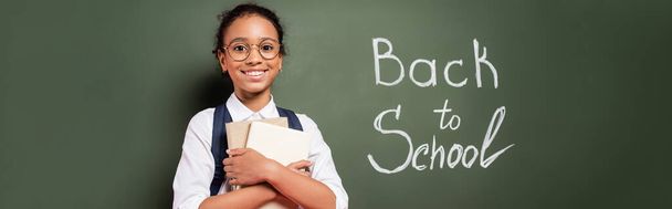 smiling african american schoolgirl with books near back to school inscription on green chalkboard, panoramic shot - Foto, Bild