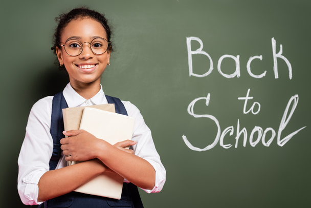 smiling african american schoolgirl with books near back to school inscription on green chalkboard - Photo, Image