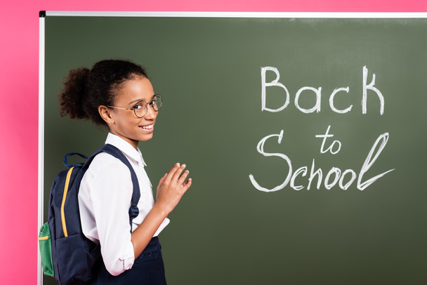 usmívající se africká americká školačka v brýlích v blízkosti zpět do školy nápis na zelené tabuli na růžovém pozadí - Fotografie, Obrázek