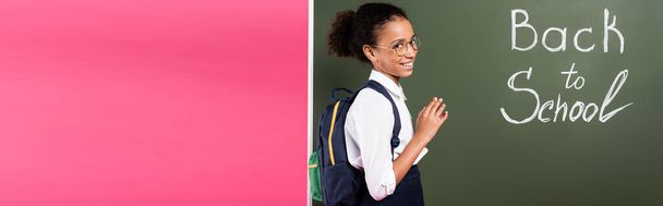 smiling african american schoolgirl in glasses near back to school inscription on green chalkboard on pink background, panoramic shot - Zdjęcie, obraz