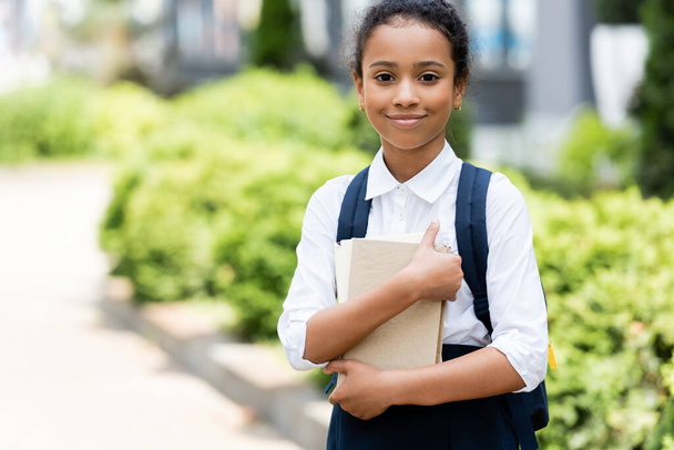 smiling african american schoolgirl with book outdoors - Photo, Image