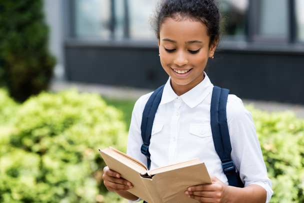 souriant afro-américaine écolière lecture livre en plein air - Photo, image