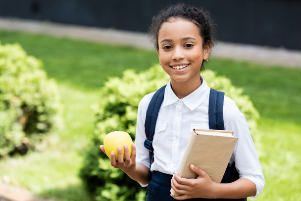 smiling african american schoolgirl with book and apple outdoors - Foto, imagen
