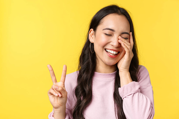 People emotions, lifestyle leisure and beauty concept. Close-up of carefree amused cute asian girl, showing peace sign touch face and laughing happy, standing yellow background - Photo, Image