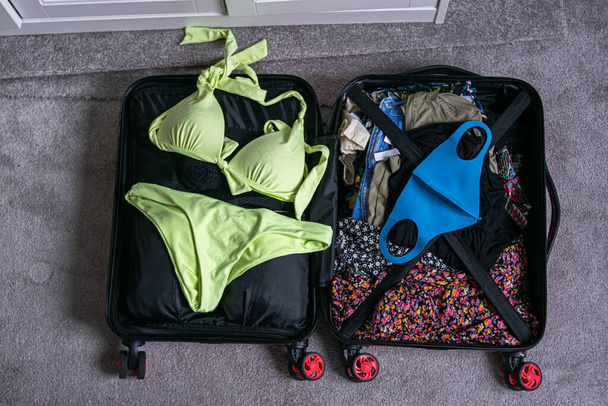 Photo d'une valise ouverte avec des vêtements d'été, un passeport et un masque réutilisable prêt à voyager.  - Photo, image