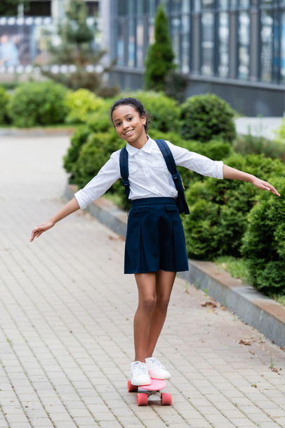 happy african american schoolgirl with backpack riding penny board outdoors - Zdjęcie, obraz