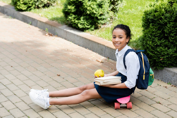 smiling african american schoolgirl with books and apple sitting on penny board - Foto, imagen