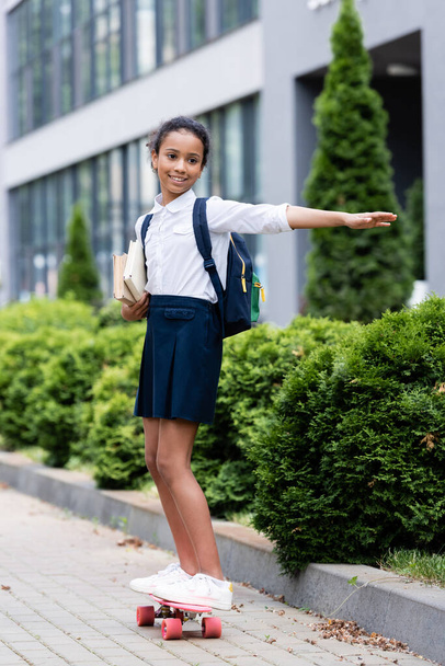 happy african american schoolgirl with backpack and books riding penny board outdoors - Photo, Image