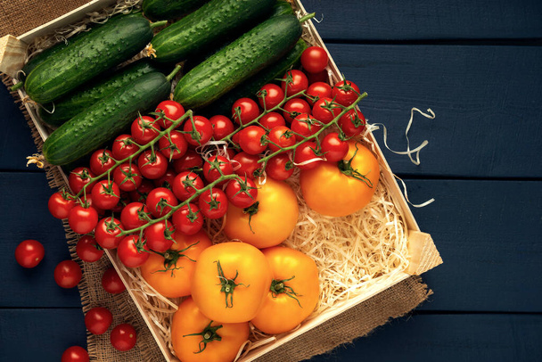 Fresh tomatoes and cucumbers in wooden box on burlap and dark wooden table. Vegetables in the box with wood shavings for transportation. Top view. Flat lay. Space for text. - Photo, Image