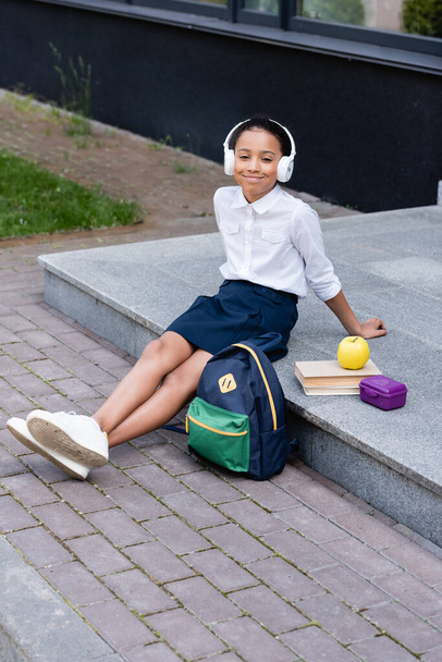 souriant afro-américaine écolière écouter de la musique dans les écouteurs près du sac à dos, livres et déjeuner en plein air - Photo, image