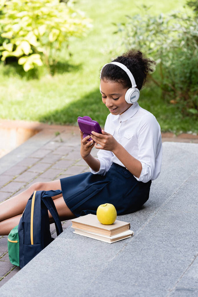 smiling african american schoolgirl listening music in headphones near backpack, books and lunch outdoors - Fotó, kép