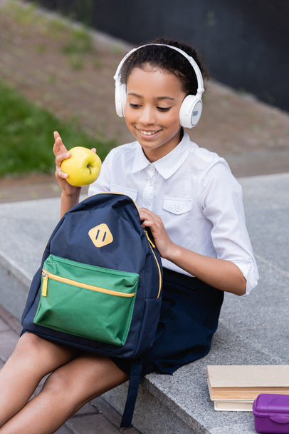 smiling african american schoolgirl in headphones holding apple and backpack near books and lunch box outdoors - Photo, Image