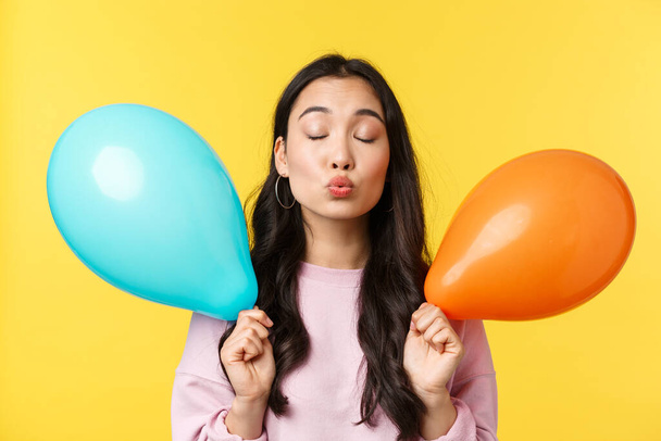 People emotions, lifestyle leisure concept. Silly and cute asian girl holding two balloons, waiting for kiss with folded lips and closed eyes, having romantic moment at party, yellow background - 写真・画像