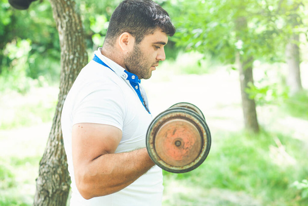Sporty man in a white t-shirt and shorts pick up heavy barbell in nature - Foto, Bild