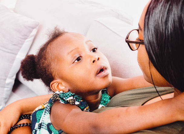 close-up van schattig klein Afrikaans Amerikaans meisje kijken naar jong mam knuffelen samen thuis. - Foto, afbeelding