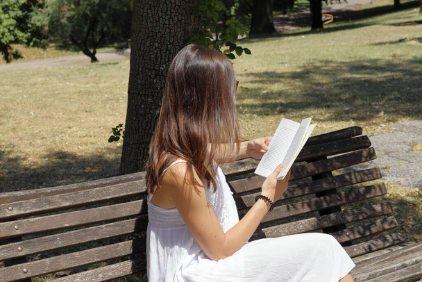 Κορίτσι με λευκό φόρεμα κάθεται σε μια τράπεζα με ένα βιβλίο - Φωτογραφία, εικόνα