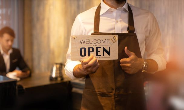 Business owner attractive young Asian man in apron hanging we're open sign on front door  welcoming clients to new cafe. Happy waiter with protective face mask holding open sign while stand at cafe . - Fotografie, Obrázek