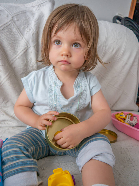 Δύο ετών γοητευτικό κοριτσάκι σε άνετα ρούχα παίζει με τα παιχνίδια της στον καναπέ στο σπίτι, με ένα ωραίο φυσικό φως. - Φωτογραφία, εικόνα