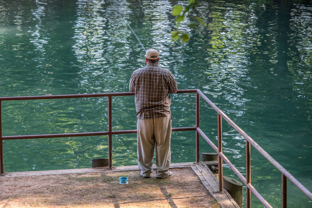 Um homem de pé no cais de pesca no lago com um pólo de pesca e um recipiente de isca ao lado dele closeup em um dia ensolarado no verão
 - Foto, Imagem