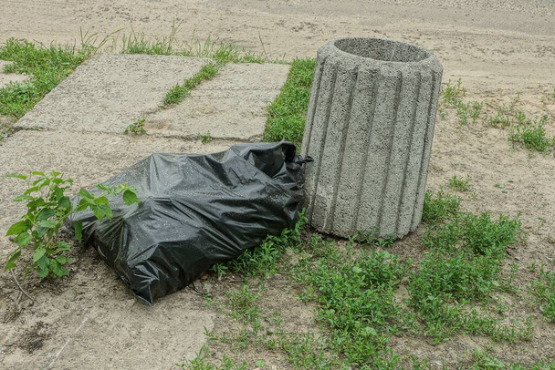 una urna de hormigón gris y una bolsa de plástico negro con basura en el suelo y hierba verde en la calle
 - Foto, imagen