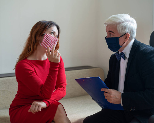 A woman in a mask at a psychologists session treats depression during an outbreak of coronavirus. A gray-haired elderly male psychologist talks to a female patient and takes notes on a clipboard. - Fotografie, Obrázek