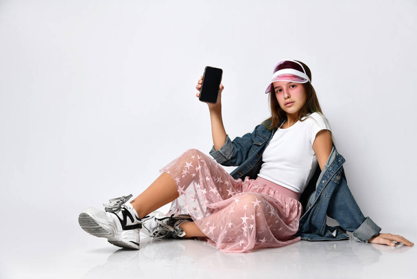 Cheerful woman sitting on the floor and showing a blank smartphone screen on a light background - Foto, immagini