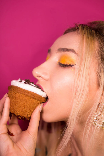 Junges Mädchen mit blonden Haaren und gelbem Make-up beißt in einen Cupcake auf rosa Hintergrund - Foto, Bild