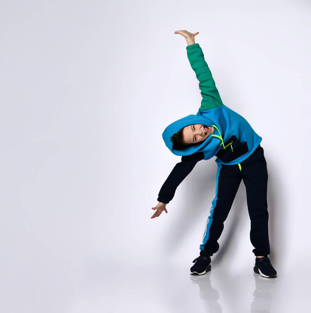 Little brunet male in hood, colorful tracksuit and black sneakers. He screaming, performing exercises, posing isolated on white - Fotó, kép