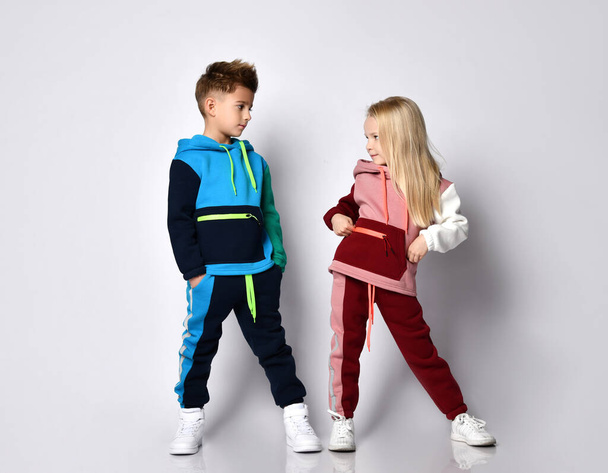 Little kids, boy and girl, in colorful tracksuits and sneakers. They posing isolated on white studio background. Hands in pockets - Φωτογραφία, εικόνα