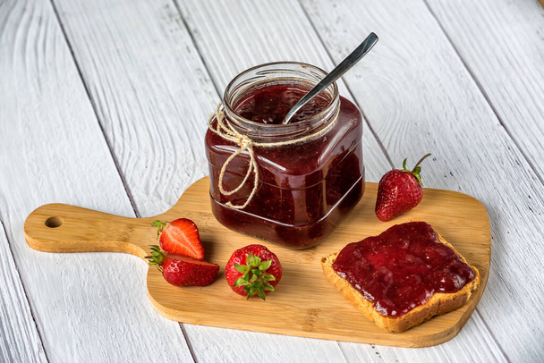 Zelfgemaakte aardbeienjam in een glazen pot en uitgespreid over een toast brood met verse aardbeien op een snijplank op een witte houten achtergrond. Rustieke stijl. - Foto, afbeelding