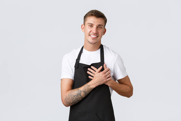 Majitelé malých podniků, kavárna a zaměstnanci koncepce. Šťastný pohledný blonďák v černé zástěře, barista nebo číšník drží ruku na srdci a usmívá se, opatrovat kavárny hosty, milovat každého zákazníka - Fotografie, Obrázek