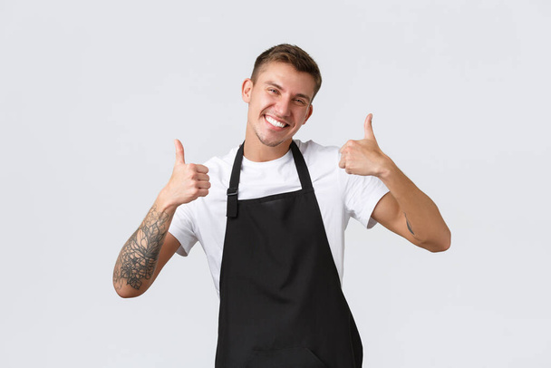 Medewerkers, kruidenierszaken en coffeeshop concept. Knappe vriendelijke barista in zwart schort, met duimen omhoog, uitnodigend voor cappuccino, glimlachend verrukt, garant voor kwaliteit, witte achtergrond - Foto, afbeelding
