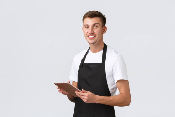 Small business, coffee shop and cafe employees concept. Handsome waiter, barista wearing apron, taking order, saving tables with digital tablet, smiling at camera, white background - Photo, Image