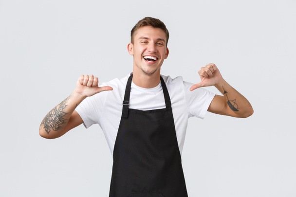 Small business owners, cafe shop and staff concept. Handsome happy barista in black apron guarantee perfect service during his shift, pointing himself and laughing joyful, make best coffee in town - Photo, Image