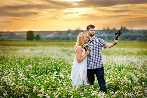 Mies ja nainen kuvaavat itseään Steadicamin avulla kamomillapellolla auringonlaskun aikaan. - Valokuva, kuva