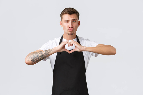 Employees, grocery stores and coffee shop concept. Cheeky and funny barista in black apron showing heart sign and pouting, blowing air kiss, love his cafe guests, inviting for drink, white background - Photo, Image