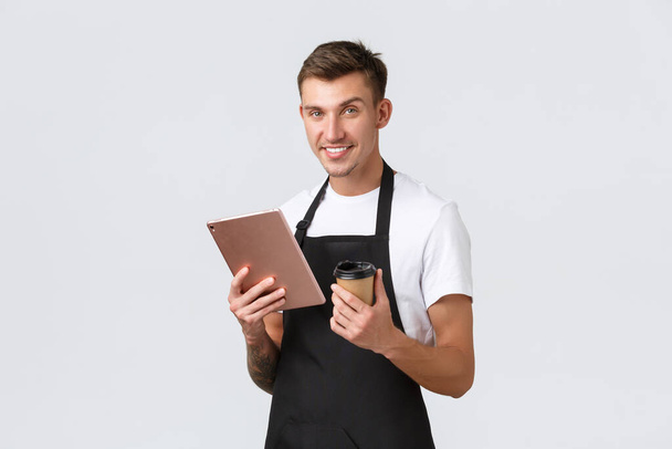 Koncepcja małych firm, kawiarni, kawiarni i restauracji. Przystojny uśmiechnięty barista, kelner w czarnym fartuchu obsługujący klienta, podający napój na wynos i trzymający cyfrowy tablet, białe tło - Zdjęcie, obraz