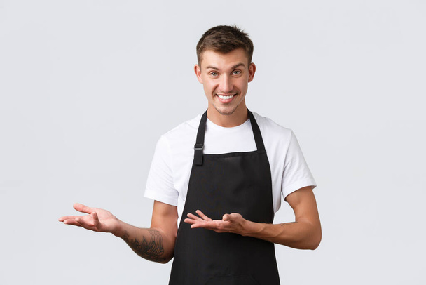Cafe and restaurants, coffee shop owners, retail concept. Handsome cheeky salesman, waiter in black apron pointing left, inviting come inside, showing way or introducing discounts, white background - Photo, Image